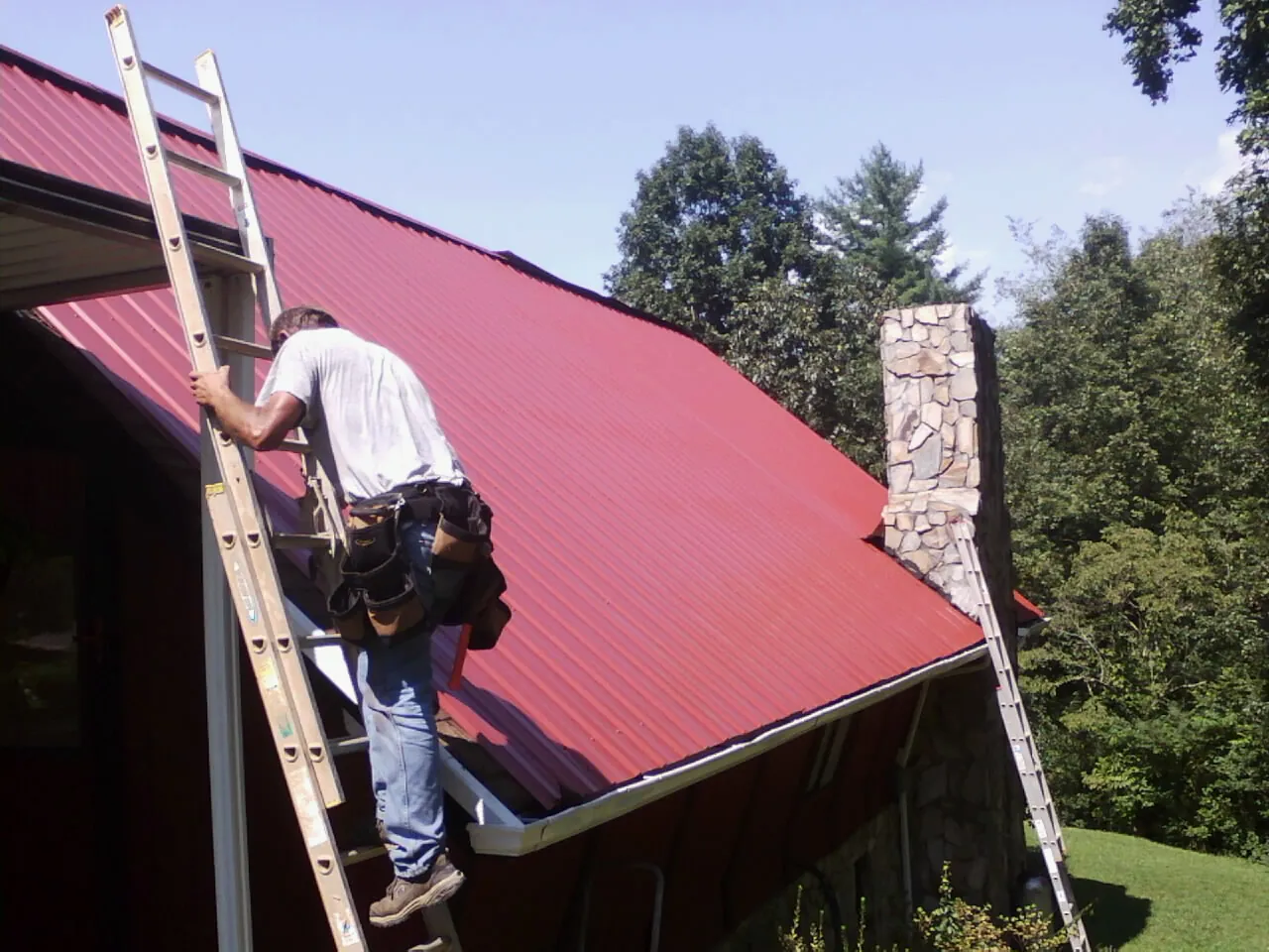 Roofer on a ladder beside a red metal roof and stone chimney — Soffit & Fascia Repair by Roofing Brothers in Somerset